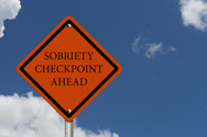 An orange diamond shaped roadsign, with the words 'Sobriety Checkpoint Ahead' in black letters.